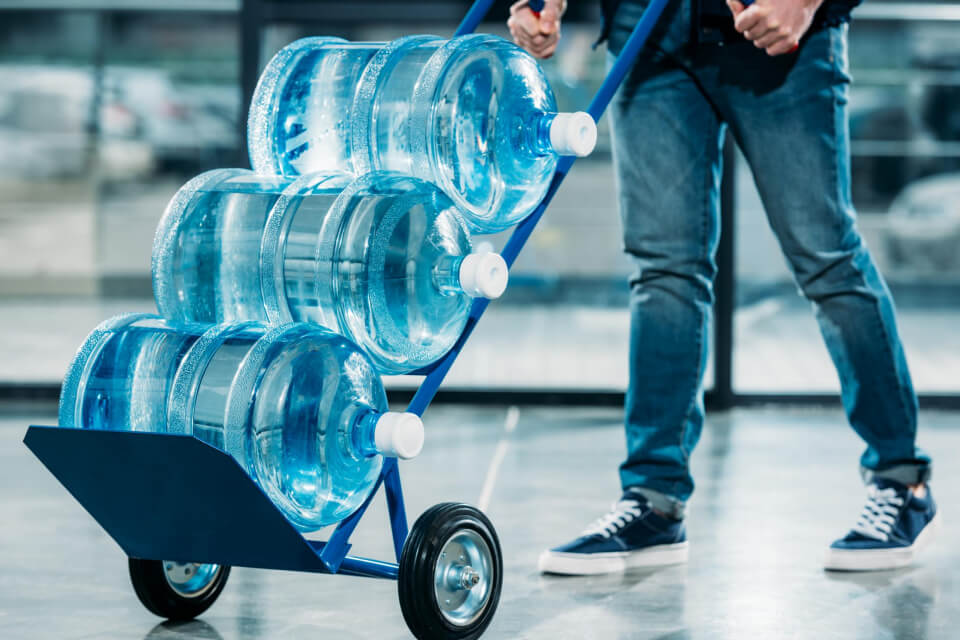 Medewerker van H2O levert grote waterflessen van 18,9 liter voor waterkoelers met een transportkar in een bedrijfsomgeving.